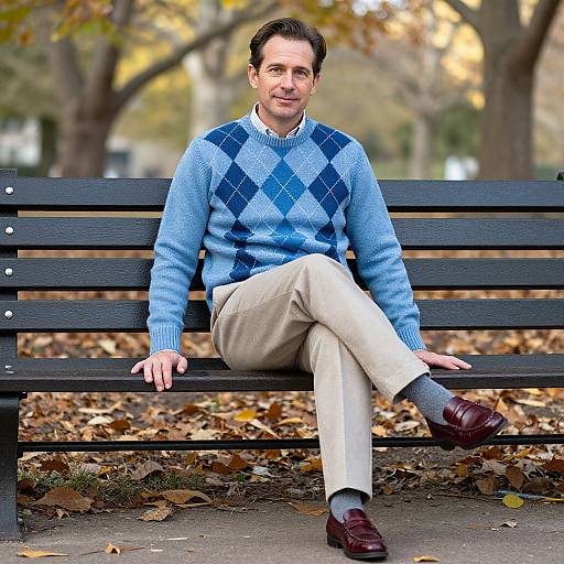 Photograph of a middle-aged man with short brown hair, wearing a blue diamond-patterned sweater, beige pants, gray socks, and brown shoes,