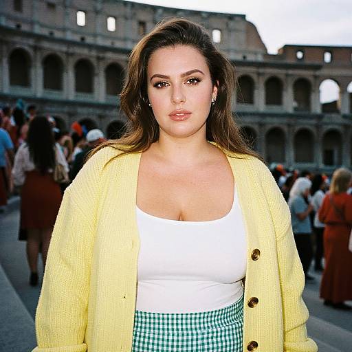 Photograph of a curvy woman with light skin, brown hair, wearing a yellow cardigan, white top, and green checkered skirt, standing