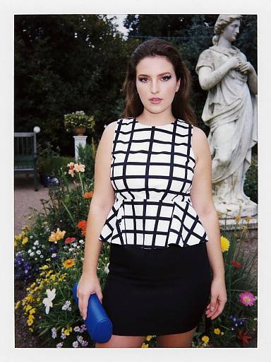 Photograph of a woman with long dark hair in a black-and-white grid pattern top and black skirt, holding a blue clutch, standing in front of