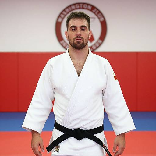 Photograph of a serious, bearded male judoka with short dark hair, wearing a white gi with black belt, standing in a red, white