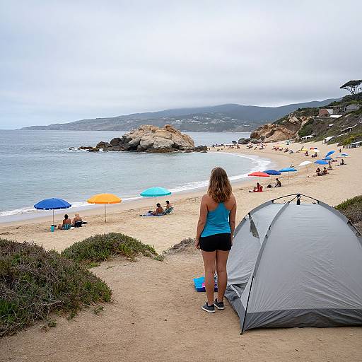 Photograph of a woman in a blue tank top and black shorts standing beside a gray tent on a sandy beach, overlooking colorful umbrellas and distant rocky