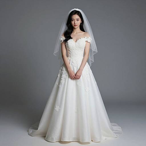 Photograph of an Asian bride in a white, off-shoulder lace wedding gown with a full skirt and veil, standing against a gray studio background