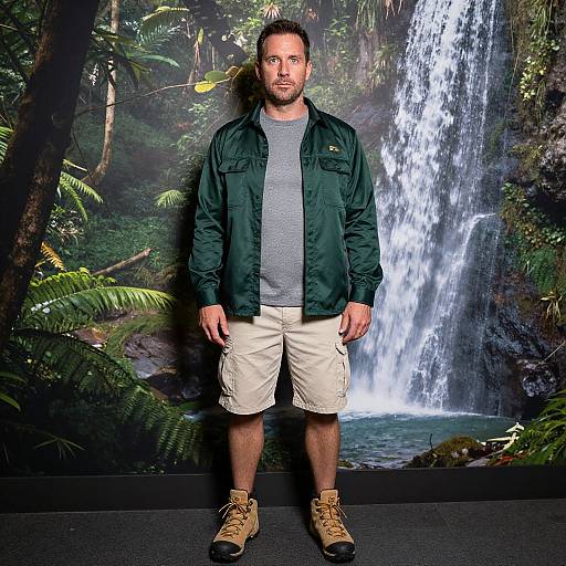 Photograph of a bearded man with short brown hair, wearing a green jacket, gray shirt, beige shorts, and tan boots, standing in front