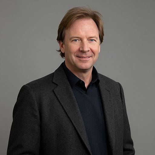 Photograph of middle-aged white man with brown hair, smiling, wearing black shirt and textured dark gray blazer, against plain gray background.
