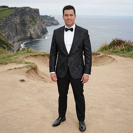 Photograph of a smiling man in a black tuxedo with bow tie, standing on a coastal cliff path, overlooking rugged cliffs and ocean.
