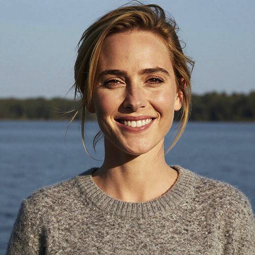 Photograph of a smiling woman with light brown hair in a loose bun, wearing a gray sweater, against a clear blue sky and calm lake background.