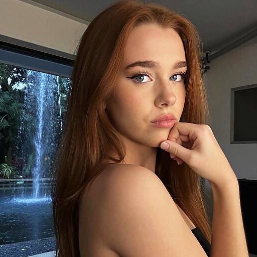 Photograph of a beautiful young woman with long brown hair, light skin, and blue eyes, posing indoors by a window with a waterfall outside. She