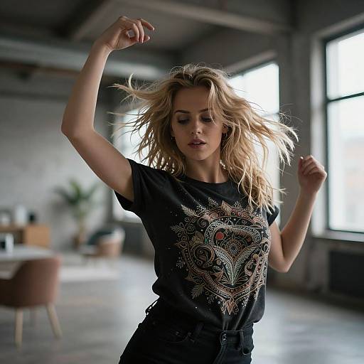 Photograph of a blonde woman with wavy hair, wearing a black T-shirt with an intricate pattern, dancing in a bright, industrial-style room with