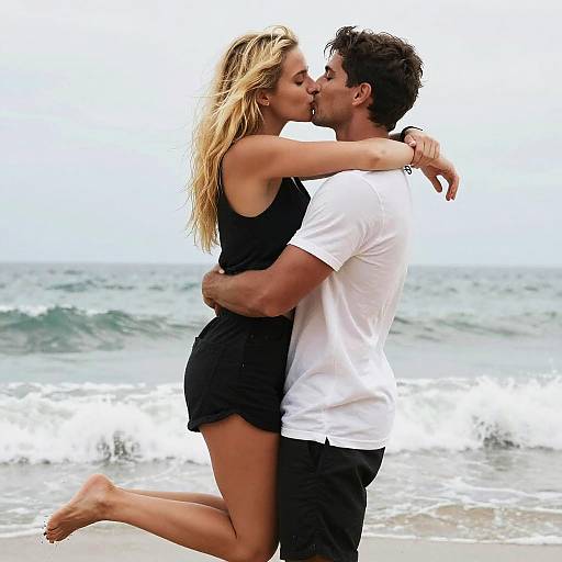 Photograph of a couple kissing on a beach; blonde woman in black dress, muscular man in white t-shirt, ocean waves in background.