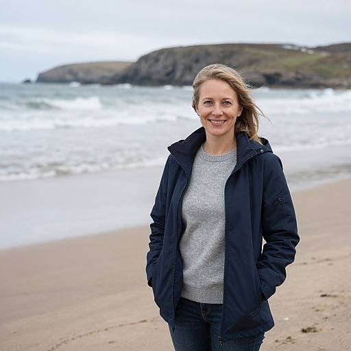 Photograph of a smiling Caucasian woman with blonde hair, wearing a black jacket and gray sweater, standing on a sandy beach with waves and rocky cliffs in