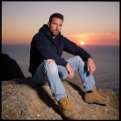 Photograph of a muscular, bearded man with short brown hair, wearing a black hoodie, ripped jeans, and tan boots, sitting on a rocky