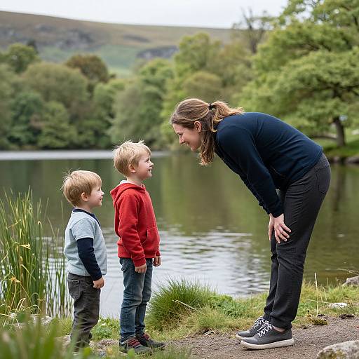 Photograph of a woman with long brown hair in a ponytail, bending down to talk to two blonde boys in red and blue hoodies by a