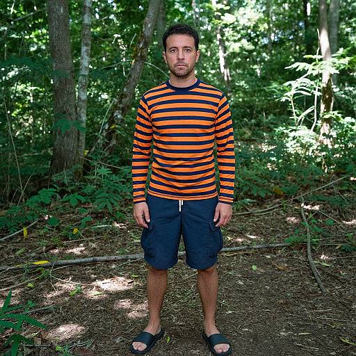 Photograph of a man with short dark hair, beard, wearing an orange and black striped long-sleeve shirt, black shorts, and sandals,