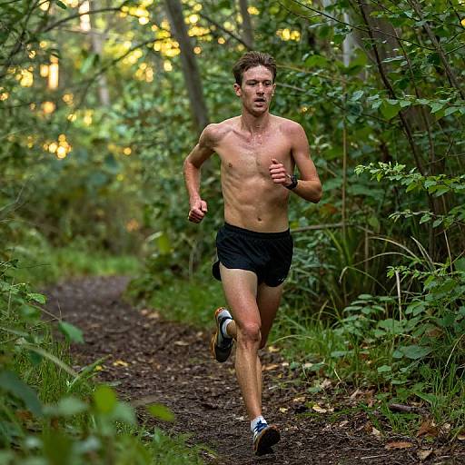 Photograph of a shirtless, slim, young white man with short brown hair jogging on a forest trail, wearing black shorts and running shoes, surrounded