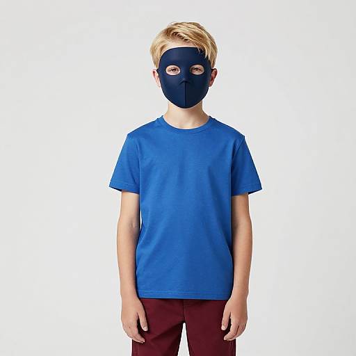 Photograph of a young boy with blonde hair, wearing a blue T-shirt, maroon pants, and a black mask with eye holes, standing against