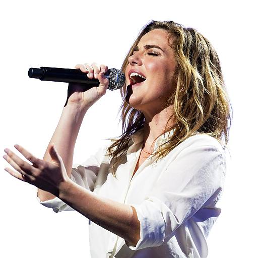 Photograph of a blonde, wavy-haired woman in a white top passionately singing into a microphone, with her hand gesturing expressively.