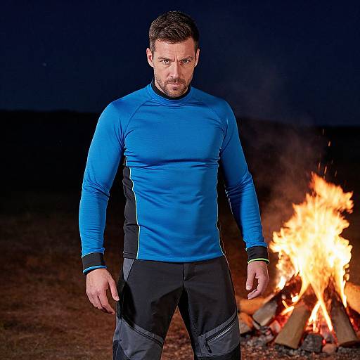 Photograph of a muscular man with short brown hair, blue long-sleeve shirt, black pants, standing by a campfire at night.