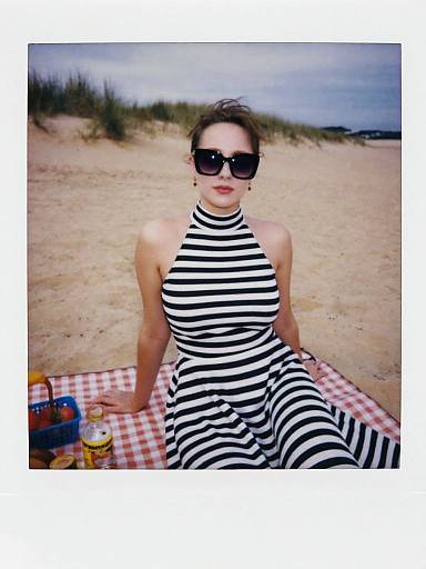 Photograph of a woman with fair skin, dark hair in a bun, wearing black-and-white striped halter dress and sunglasses, sitting on a red