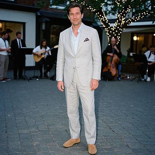 Photograph of a handsome man in a white suit, light brown shoes, and patterned pocket square, standing in an evening outdoor event with string lights