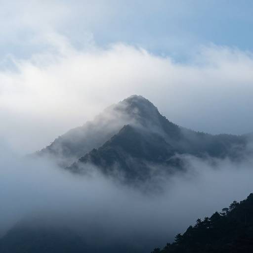 Photograph of mist-covered, dark green mountain peak with dense fog enveloping lower slopes under a bright, partly cloudy sky.