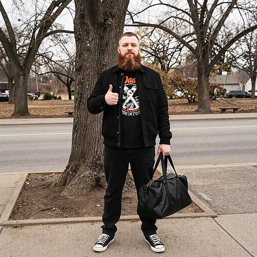 Photograph of a bearded man with a full red beard, wearing a black jacket, graphic tee, and black pants, thumbs up, holding a