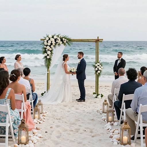 Photograph of beach wedding: bride in white gown, groom in navy suit, exchanging vows under wooden arch with white flowers, surrounded by guests seated on