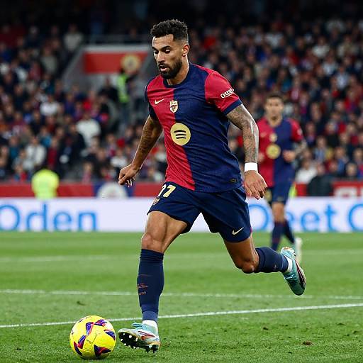 Photograph of a muscular male soccer player in a red and navy Barcelona uniform, mid-action, chasing a yellow and blue ball on a green field with