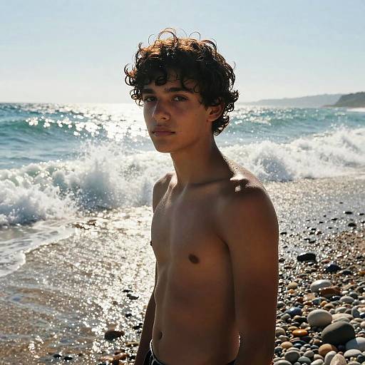 Photograph of a young, shirtless, curly-haired boy standing on a rocky beach with sparkling ocean waves and bright sunlight behind.