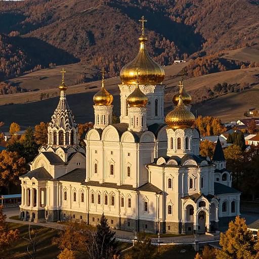 Photograph of a sunlit Orthodox church with golden domes, white walls, and intricate architectural details, surrounded by autumnal hills.