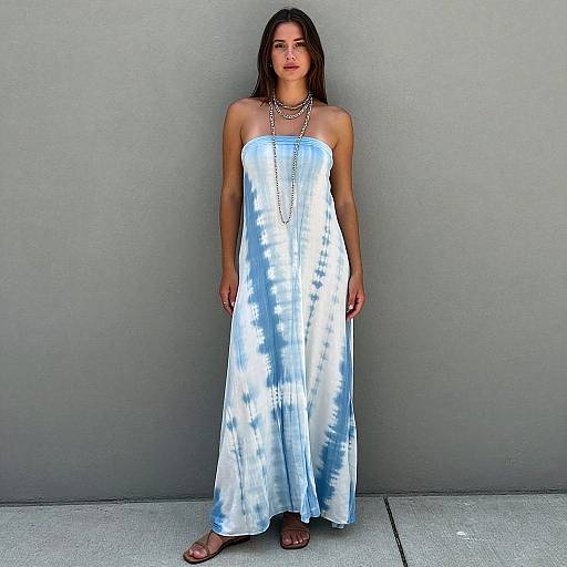 Photograph of a young woman with shoulder-length brown hair, wearing a strapless blue and white tie-dye maxi dress, long necklace, and sandals