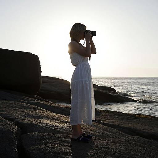 Silhouetted photograph of a woman in a white dress, holding a camera, standing on rocky shore at sunset, ocean in background.