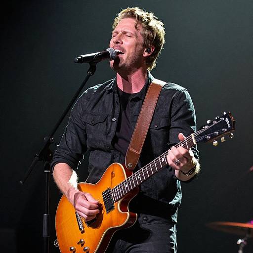Photograph of a bearded male guitarist with short blonde hair, wearing a black shirt, passionately singing into a microphone while playing an orange electric guitar on