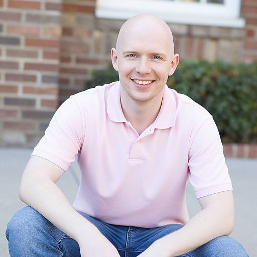 Photograph of a bald, smiling man with fair skin, wearing a light pink polo shirt and blue jeans, sitting outdoors against a brick wall.