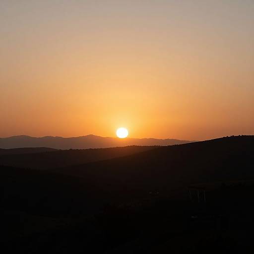 Photograph of a vibrant sunset with a glowing orange sun setting over dark, silhouetted hills and a gradient sky from orange to pale yellow.