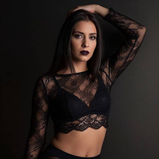 Photograph of a dark-haired woman with black lace crop top, revealing black bra, arm raised, dark lipstick, against a black background.