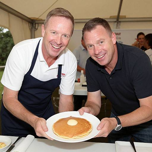 Photograph of two smiling middle-aged men, one in a white shirt and black apron, the other in a black polo, holding a pancake