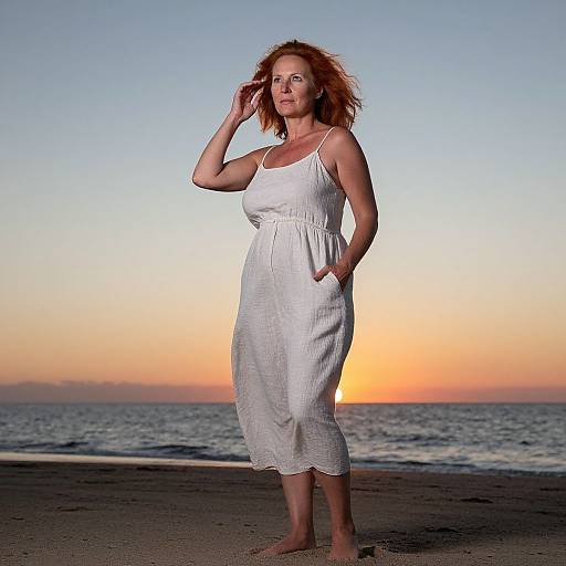 Photograph of a red-haired woman in a white sundress standing barefoot on a beach at sunset, hand in hair, ocean and orange sky in