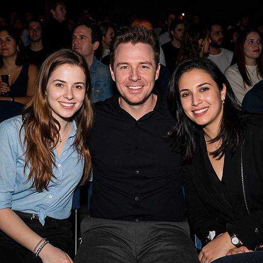 Photograph of three smiling people, man in black shirt center, woman in blue shirt left, woman in black top right, seated in dimly lit