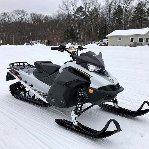 Photograph of a black and white snowmobile with orange accents parked on a snowy driveway, with a house and leafless trees in the background.