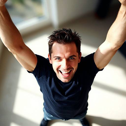 Photograph of a smiling, short-haired man with fair skin, wearing a black t-shirt, arms raised, looking up at the camera in bright,