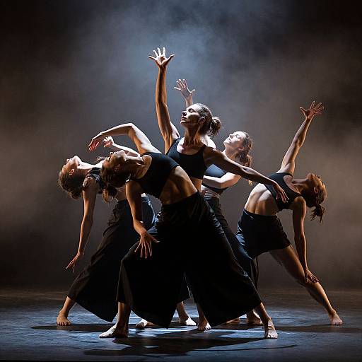 Photograph of five women in black dance attire performing a dynamic, synchronized dance with arms raised, illuminated by dramatic stage lighting.