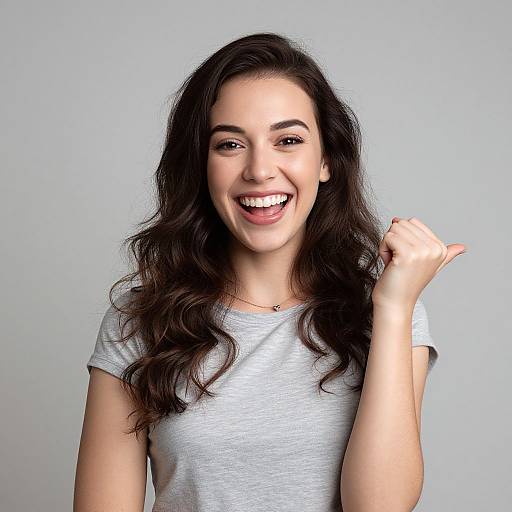 Photograph of a smiling young woman with long, wavy dark brown hair, wearing a light gray t-shirt, against a plain gray background, raising