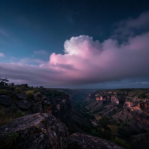 Photograph of a dramatic canyon at dusk, featuring dark rocky cliffs, a glowing pink and white cloud-streaked sky, and starry night above
