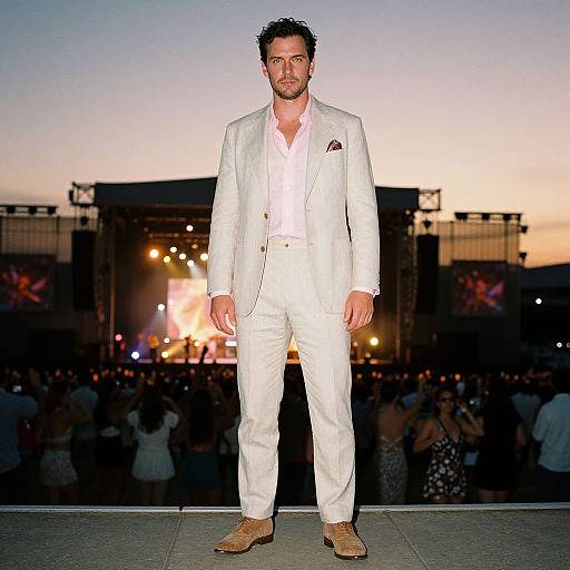 Photograph of a man in a white suit, white shirt, and brown shoes, standing on a rooftop at sunset, with a crowded background and illuminated