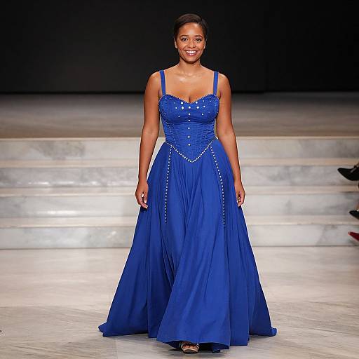Photograph of a smiling, dark-skinned woman in a vibrant blue, strapless, floor-length gown with pearl detailing, walking down a marble runway