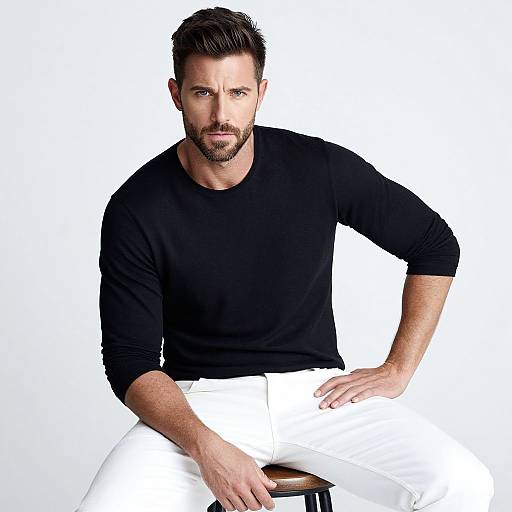 Photograph of a muscular, bearded man with short brown hair, wearing a black long-sleeve shirt and white pants, seated against a white
