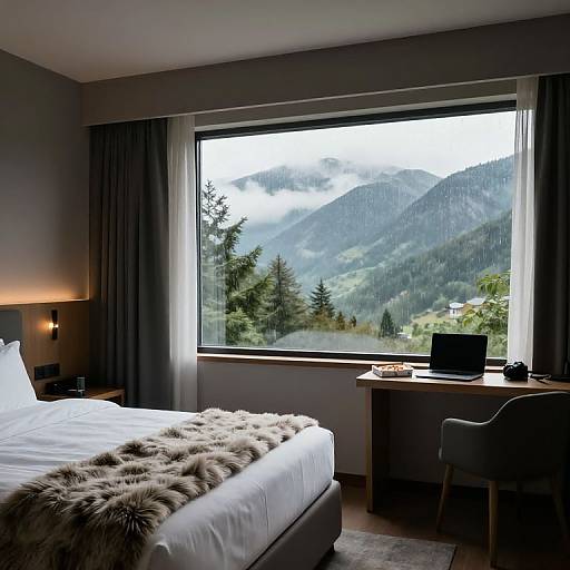 Photograph of a modern, mountain-view bedroom with a large window, white bed with fur blanket, laptop on desk, grey curtains, and a forest