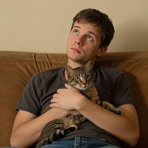 Photograph of a young white man with short brown hair, wearing a black t-shirt, sitting on a brown couch, holding a tabby cat against