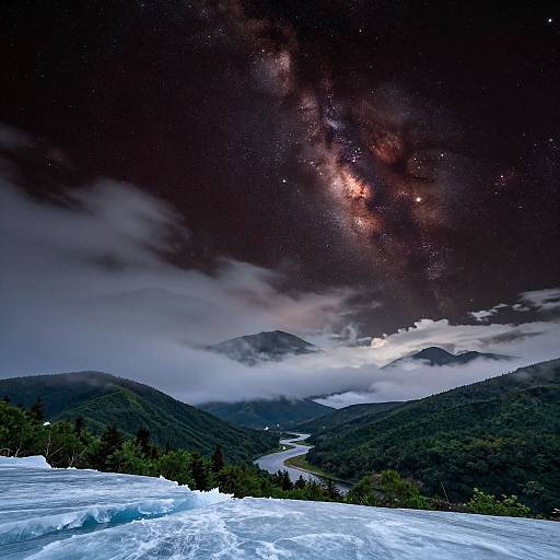 Photograph of a nighttime mountain landscape with a glowing Milky Way, misty mountains, and a river winding through dense forest.