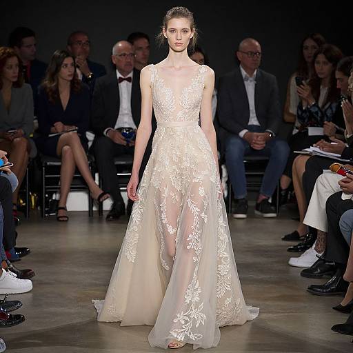 Photograph of a slender, pale-skinned female model in a sheer, white lace bridal gown, walking down a dimly lit runway with seated audience